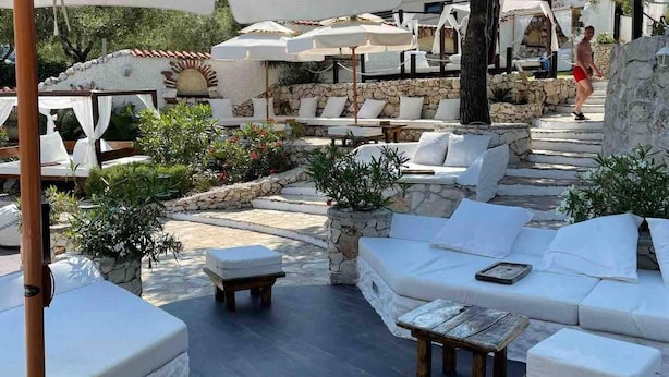A sunlit outdoor seating area with stone walls, white cushions, and wooden tables. - Cozy Loft Studio With Rooftop, Seaview & Pool (Okrug)