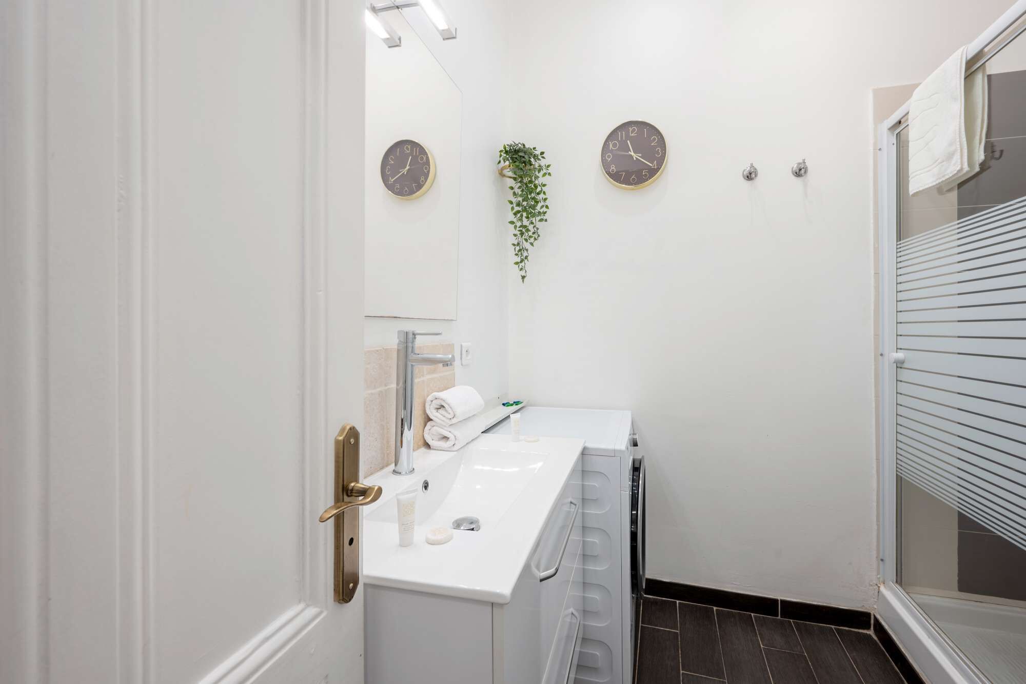 A modern bathroom with a white sink, two wall clocks, a towel rack, and a small potted plant.