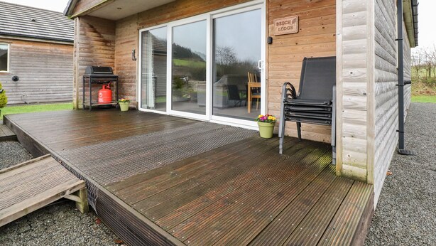 Un chalet en bois doté d’un patio couvert, d’un barbecue et d’un espace détente avec une chaise et des plantes en pot.