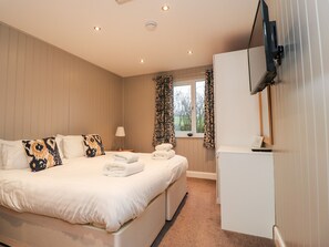 A neatly made bed with white linens and decorative pillows, a bedside table with a lamp, a window with curtains, and a television mounted on the wall. - Beech Lodge (Preston)