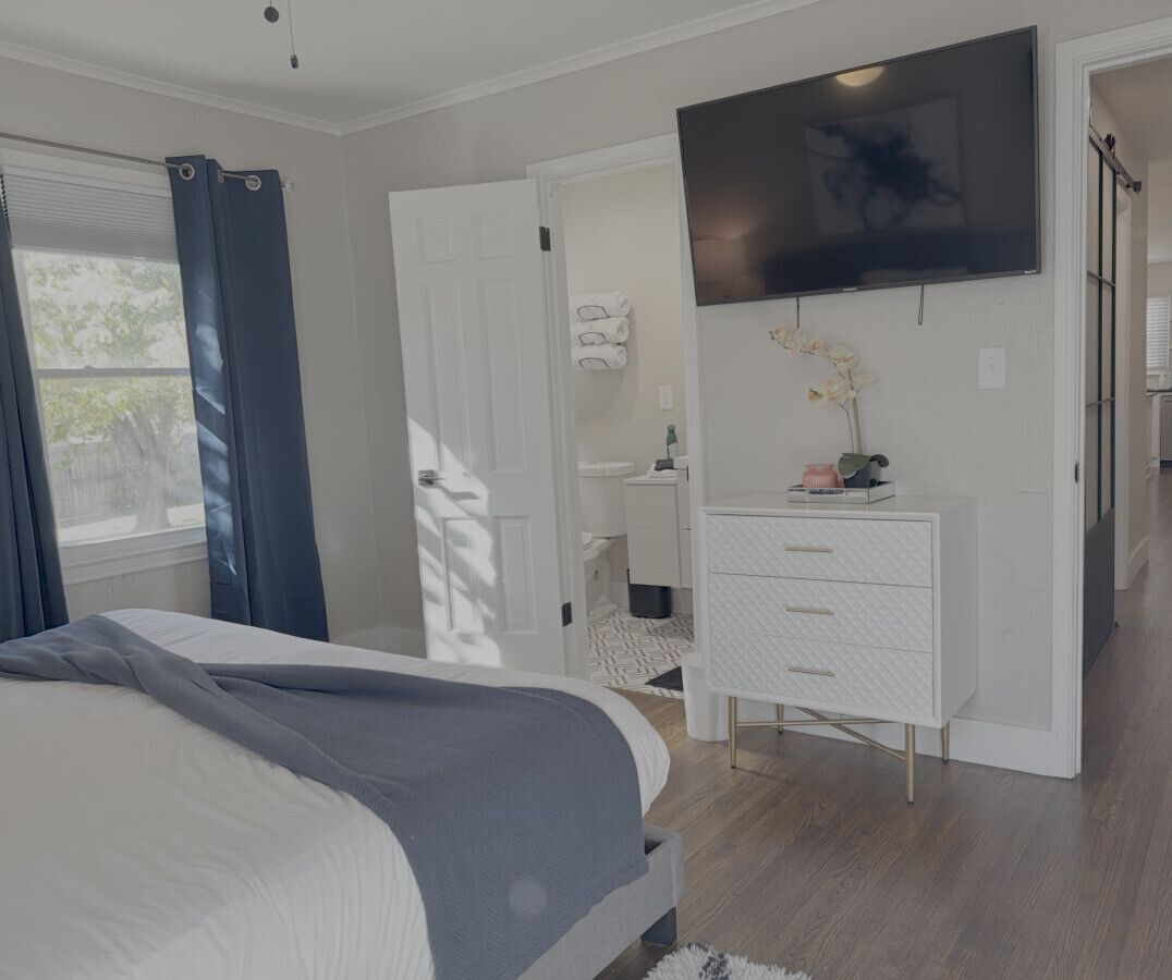 Un dormitorio con cama, televisor montado en la pared, cómoda y baño visible a través de una puerta abierta.
