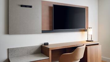 A modern hotel room with a wooden desk, a flat-screen TV mounted on the wall, a lamp, and a cushioned bench.