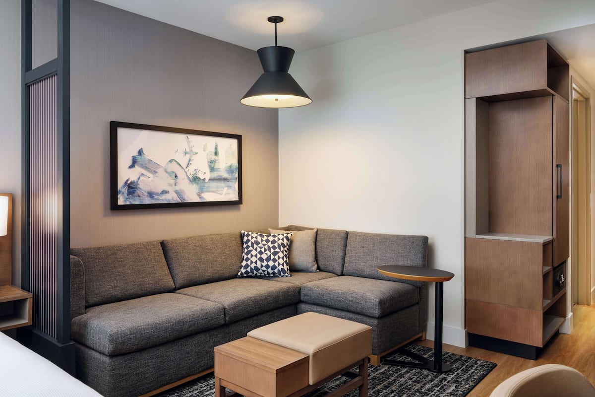 Een moderne woonkamer met een hoekbank, een bijzettafeltje, een klein houten bankje, een lijstwerk abstract schilderij en een ingebouwde houten boekenkast.