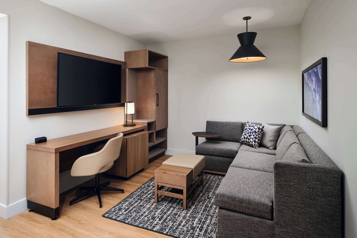 Een moderne woonkamer met een houten bureau, een grijze bank, een zwarte hanglamp en een ingelijst schilderij aan de muur.