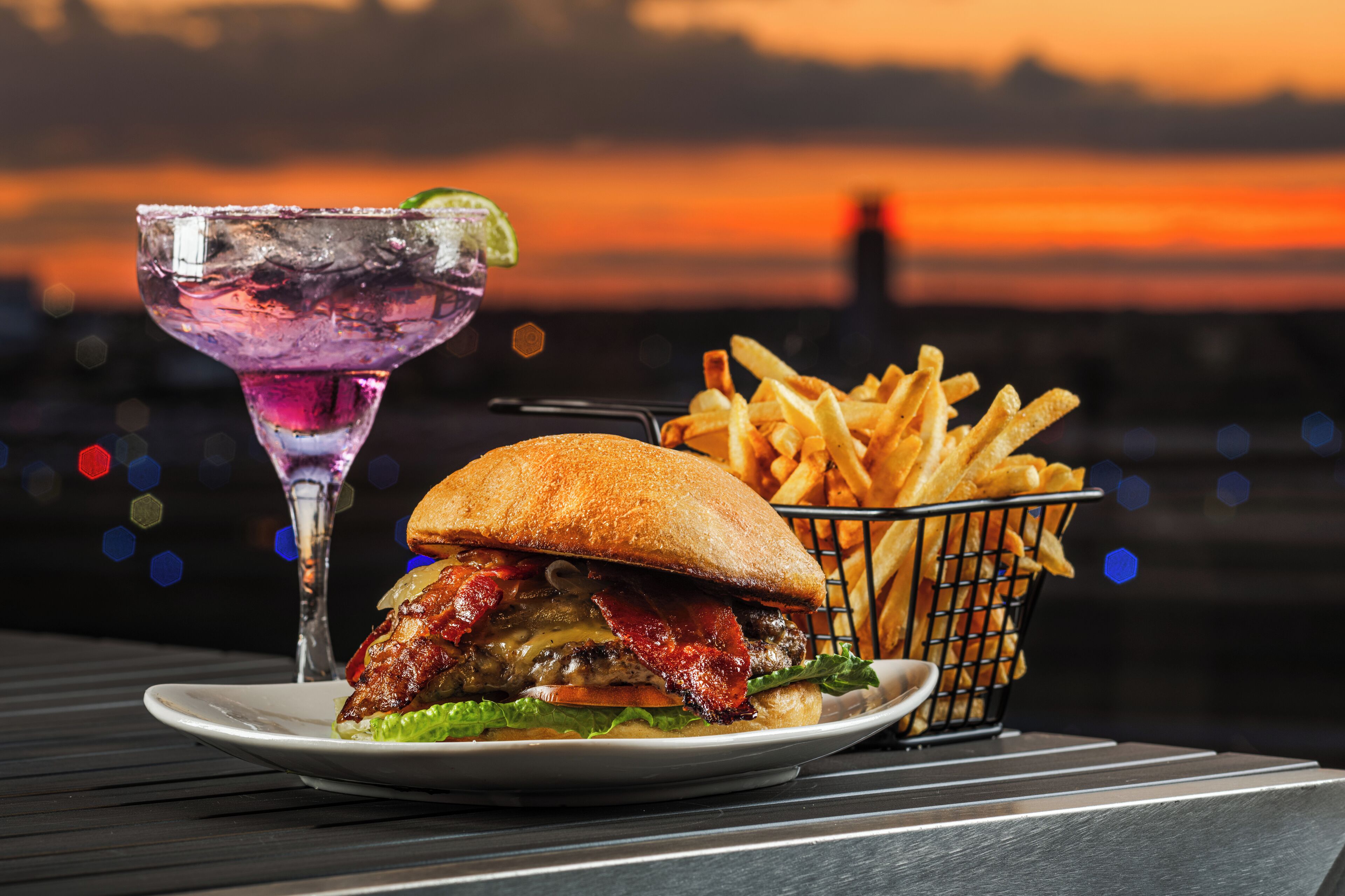 A burger with fries and a cocktail on a table with a sunset view.
