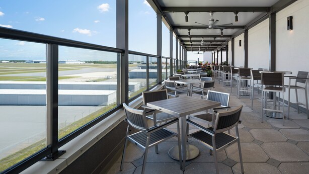 Een overdekt buitenterrein met tafels en stoelen, grote ramen met uitzicht op een landingsbaan, en een blik op het vliegveld.