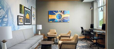 A modern hotel room with a sofa, armchairs, a coffee table, and a desk with computers. There are framed artworks on the wall and a large window providing natural light.
