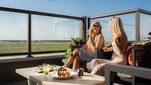 Een moderne kamer met grote ramen en uitzicht op een luchthaven. Er staat een tafel met eten en drinken, en twee personen zitten op een bank.