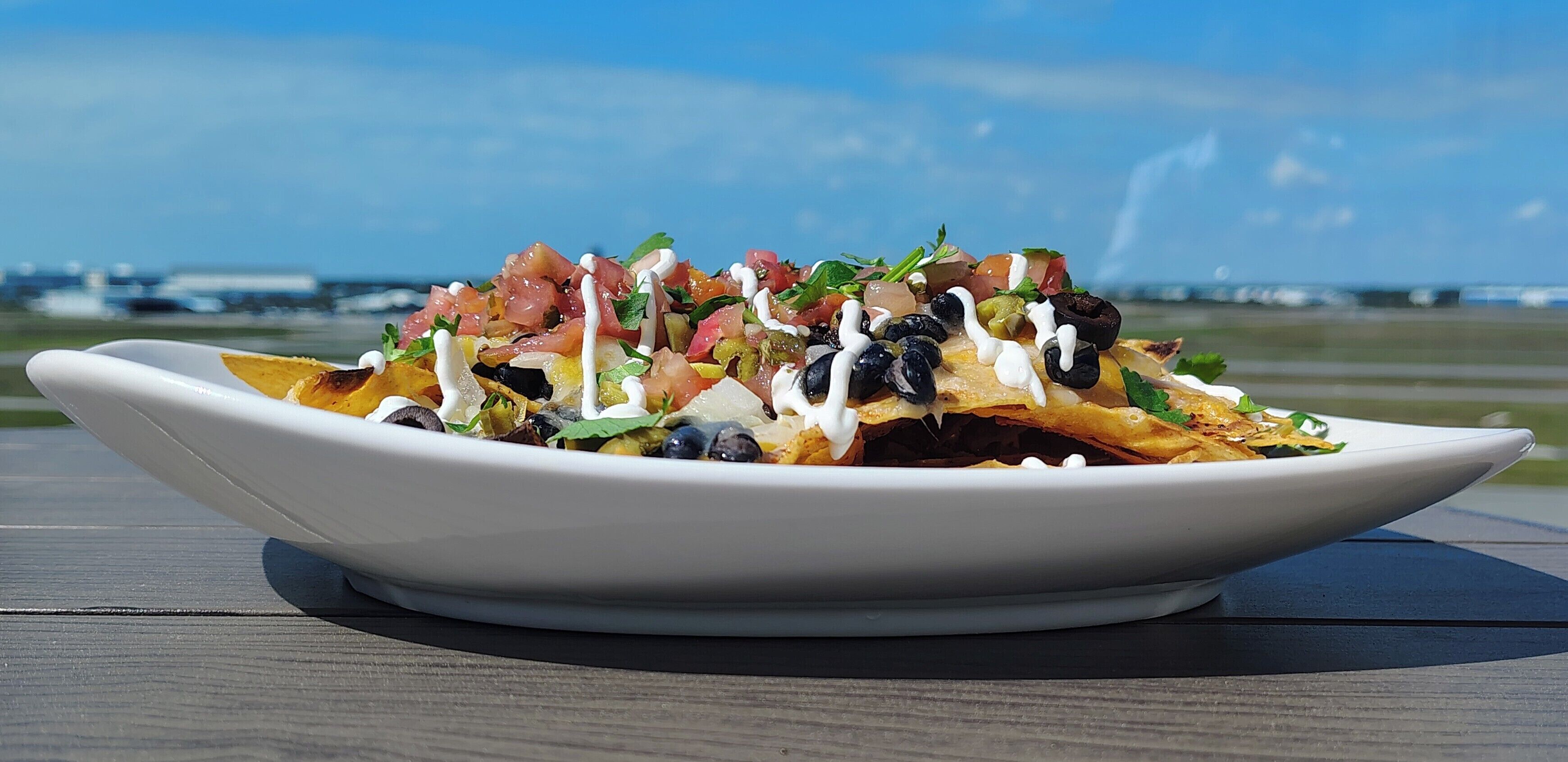 Nachos on a white plate with various toppings, placed on a wooden table with a clear sky and distant airport in the background.