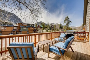 Una terraza de madera con mobiliario exterior, una parrilla y vistas a casas de montaña.