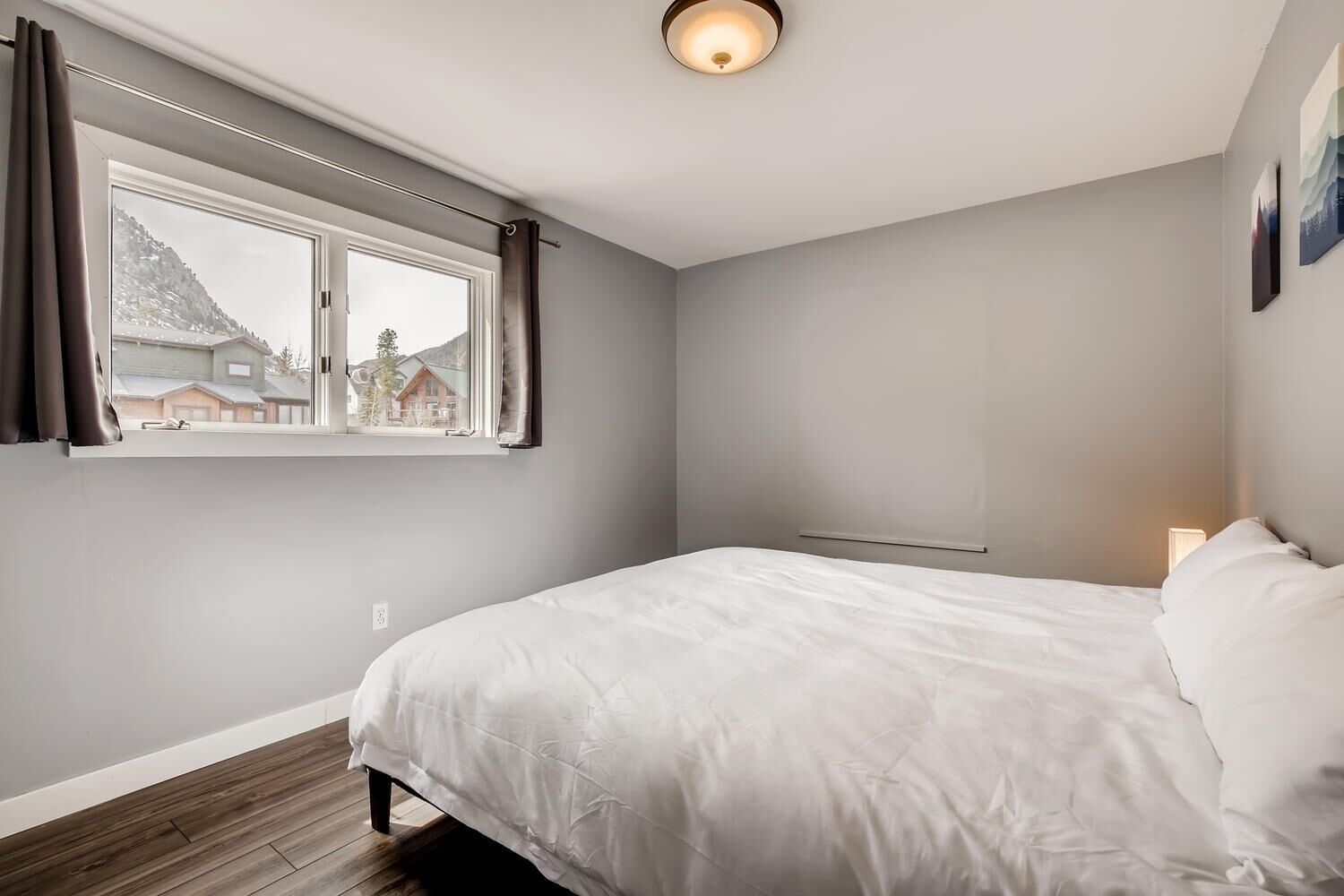 A bedroom with a large bed, a window with curtains, a bedside table with a lamp, and a view of houses and mountains.
