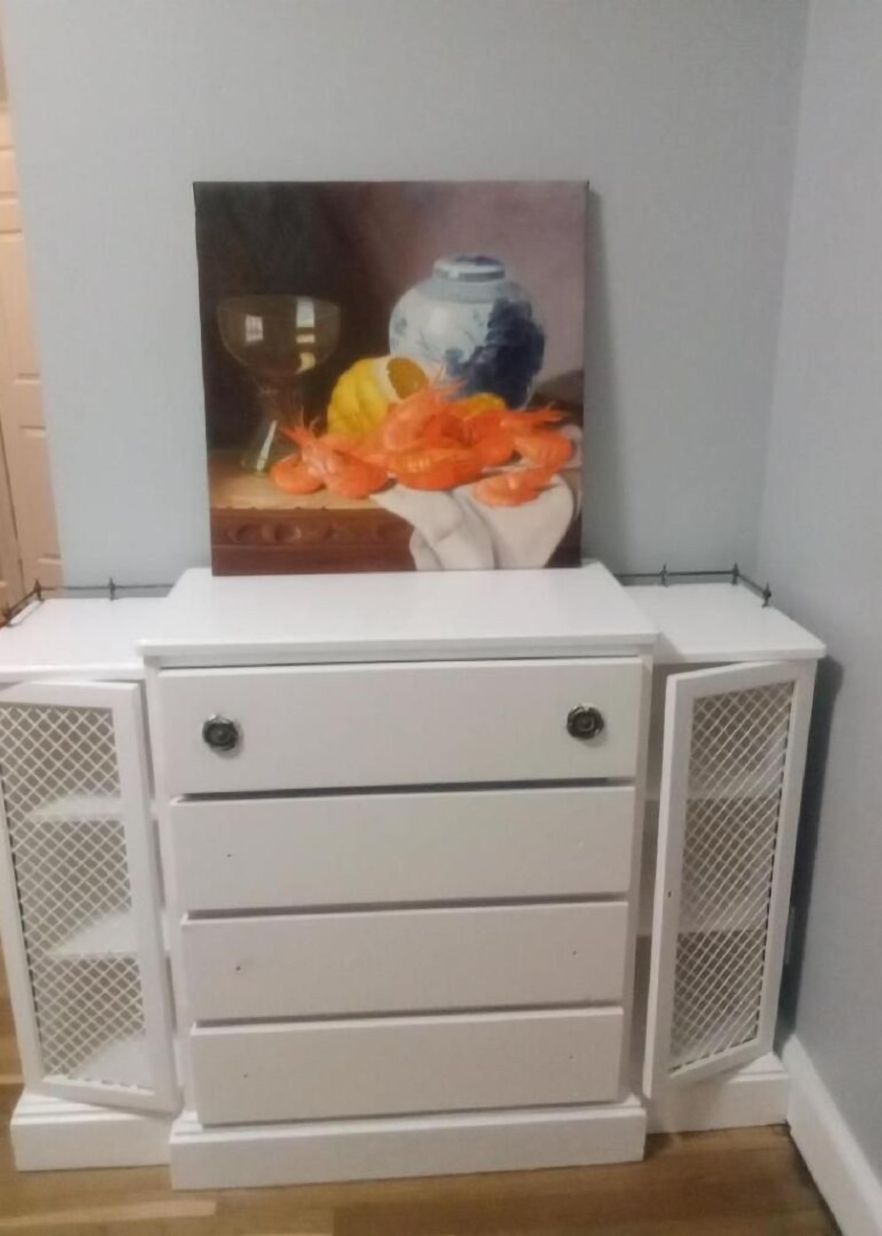 Ein weißer Nachttisch mit drei Schubladen und zwei Schrankelementen mit Mesh-Geflecht, an einer Wand.
