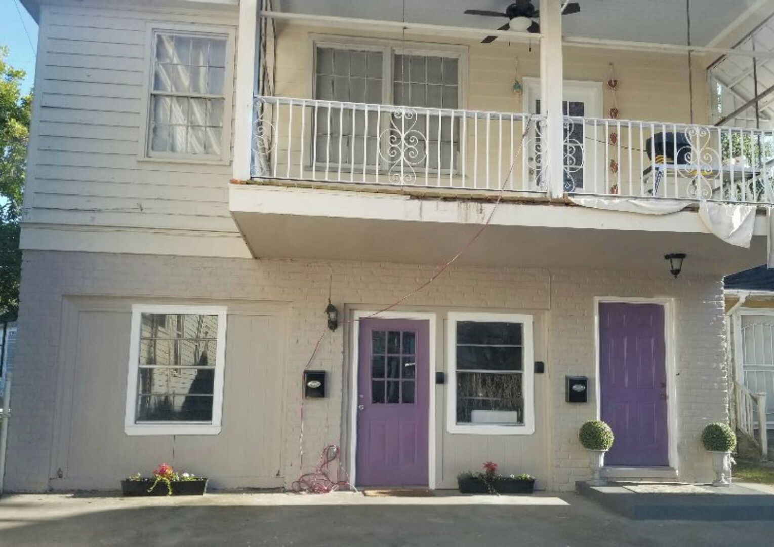 A two-story building with a balcony, a purple door, and a small porch area with potted plants.