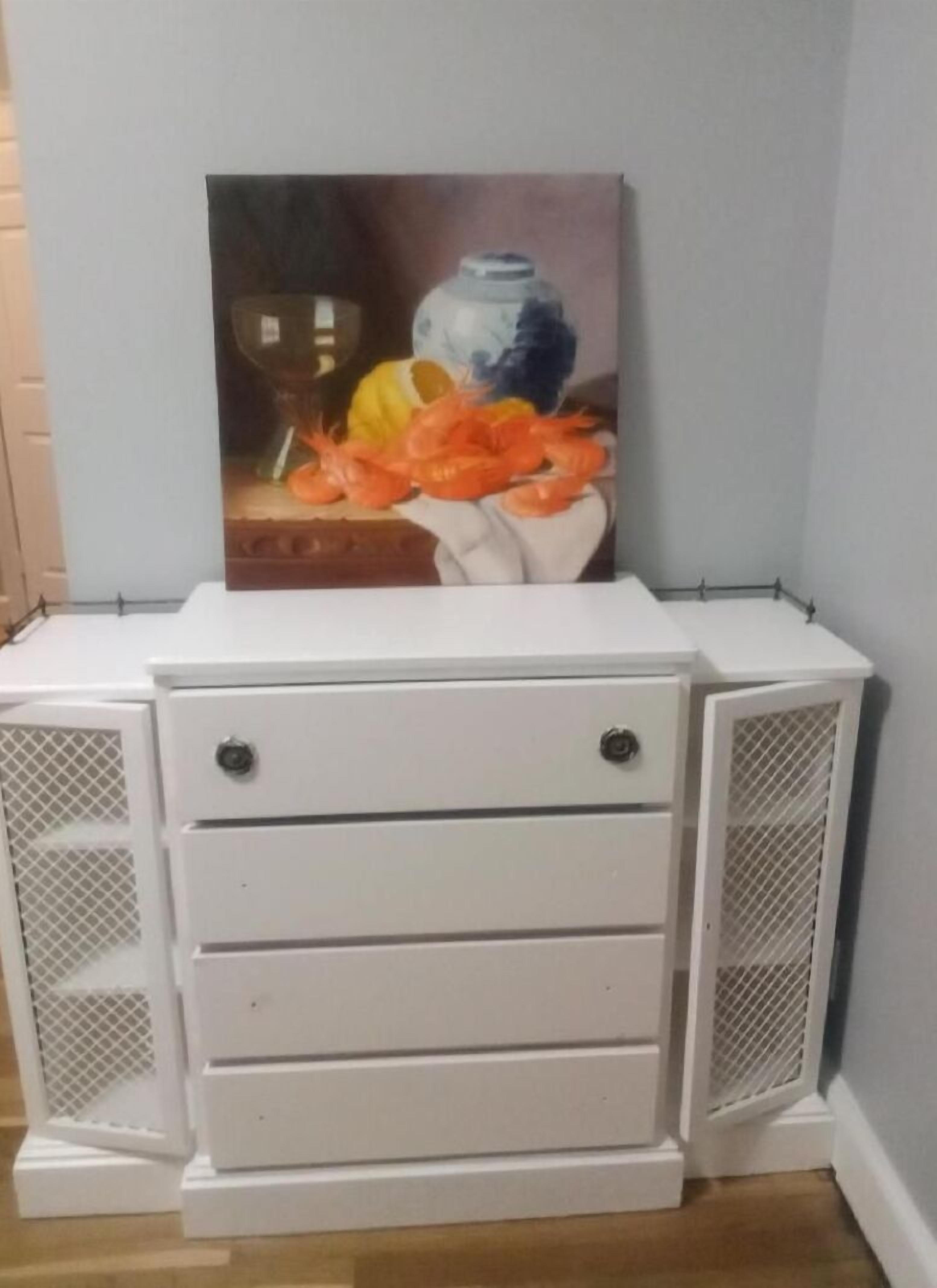 A white bedside table with three drawers and two mesh-fronted cabinets against a wall.