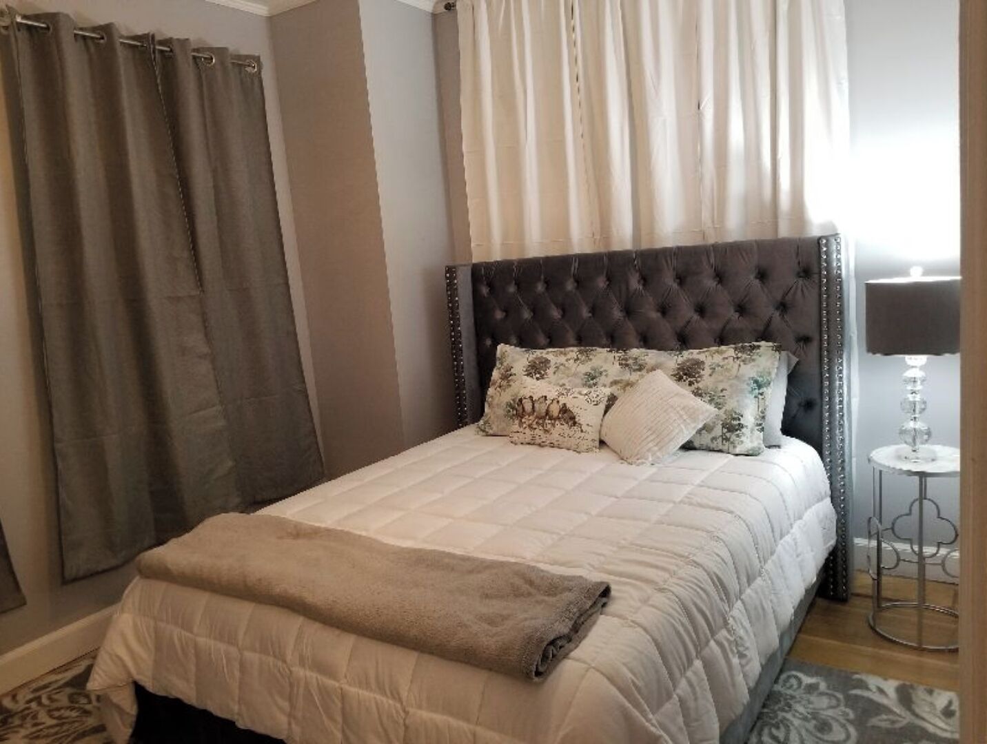 Una cama bien hecha con cabecera capitonada, una lámpara de noche y una alfombra estampada.