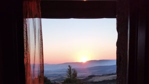 Une vue à travers une fenêtre ornée de rideaux légers, donnant sur un champ verdoyant et des collines lointaines au coucher du soleil.