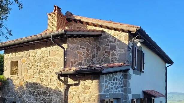 Une maison en pierre avec un toit en tuiles, un petit balcon et des plantes en pot disposées autour de l’entrée.