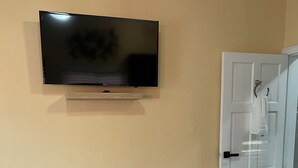 Une chambre à coucher avec un lit, un fauteuil gris, un téléviseur fixé au mur et une porte blanche sur laquelle est accrochée une serviette.