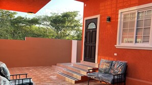 Une terrasse couverte avec un sol carrelé, un escalier menant à une porte et deux chaises recouvertes de housses en plastique.