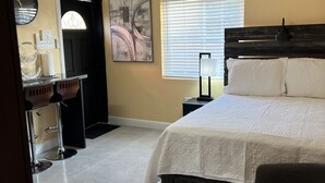 Une chambre à coucher comprenant un lit, une table de chevet, une lampe, un appareil de climatisation et une fenêtre avec des stores.