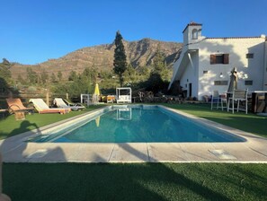 Una piscina con sillas de descanso y un edificio blanco con una torre de campanas, todo ello rodeado de montañas.