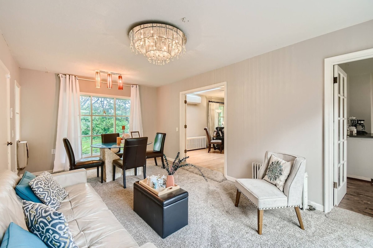Ein Wohnzimmer mit einer Couch, Hocker und Sesseln, ein Essbereich mit Stühlen und einem Tisch, sowie eine Deckenleuchte.