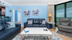 Un salon avec un canapé bleu, un fauteuil gris et une table basse. Deux tableaux abstraits ornent le mur, ainsi qu’une lampe. Une grande fenêtre offre une vue sur l’extérieur.