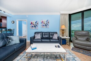 A living room with a blue couch, a grey armchair, and a coffee table. There are two abstract paintings on the wall, a lamp, and a view of the outdoors through a large window.