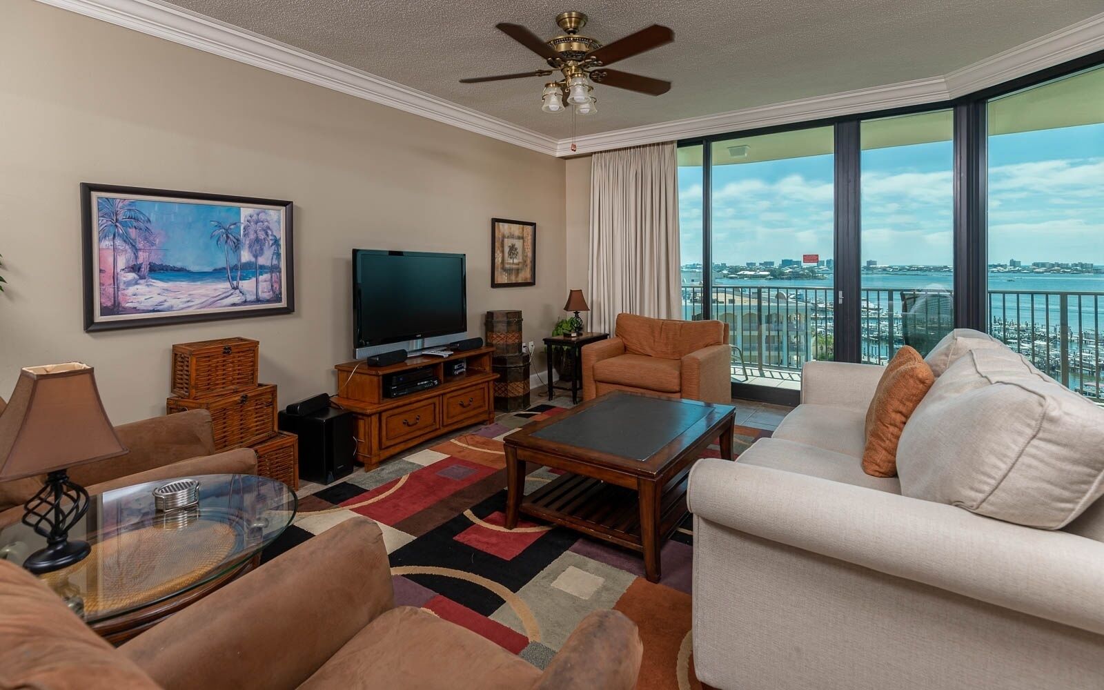 A spacious living room with a large flat-screen TV, comfortable seating, a coffee table, and a balcony with a city view.