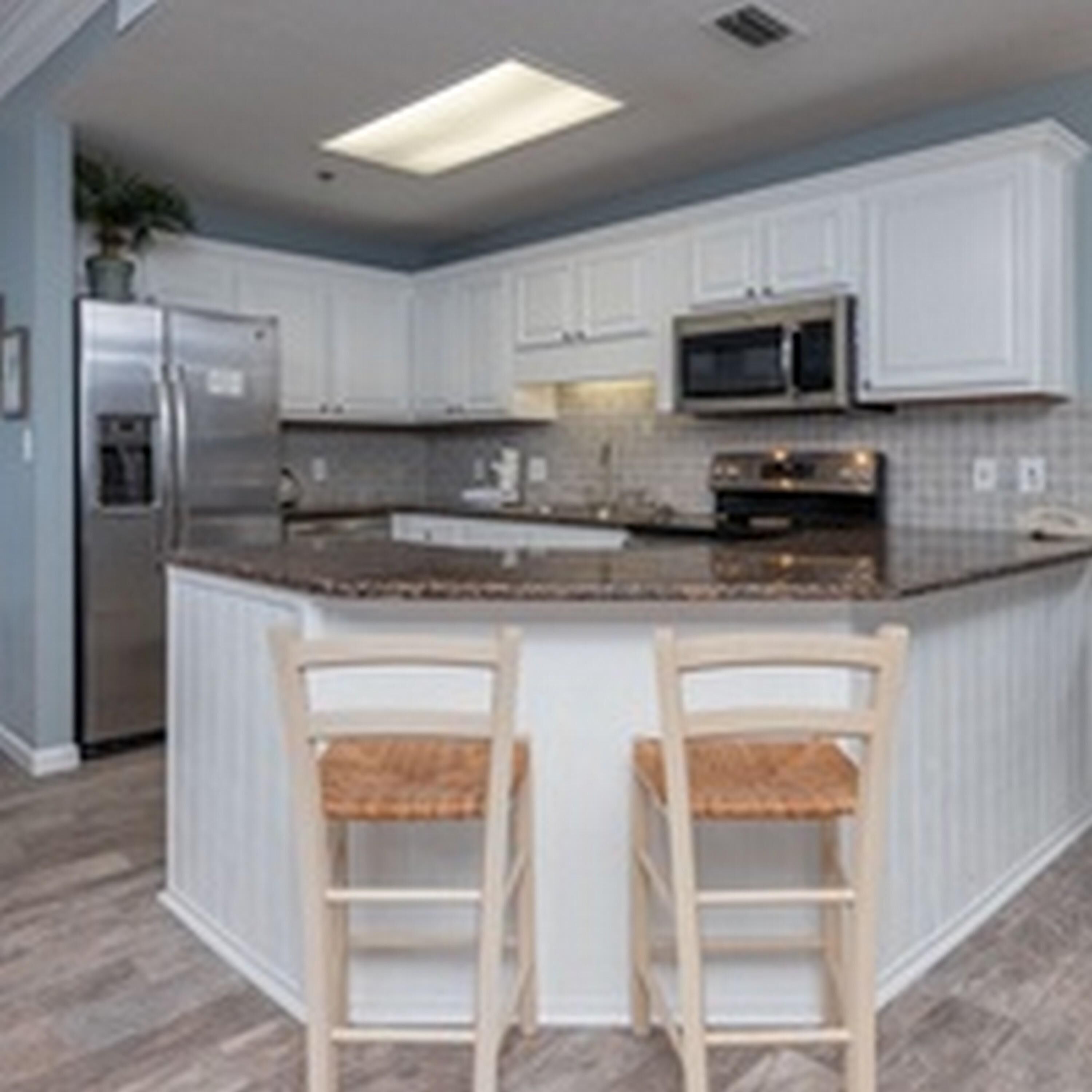A kitchen with white cabinets, a stainless steel refrigerator, a microwave, and a granite countertop. There are two wooden bar stools with cushioned seats.