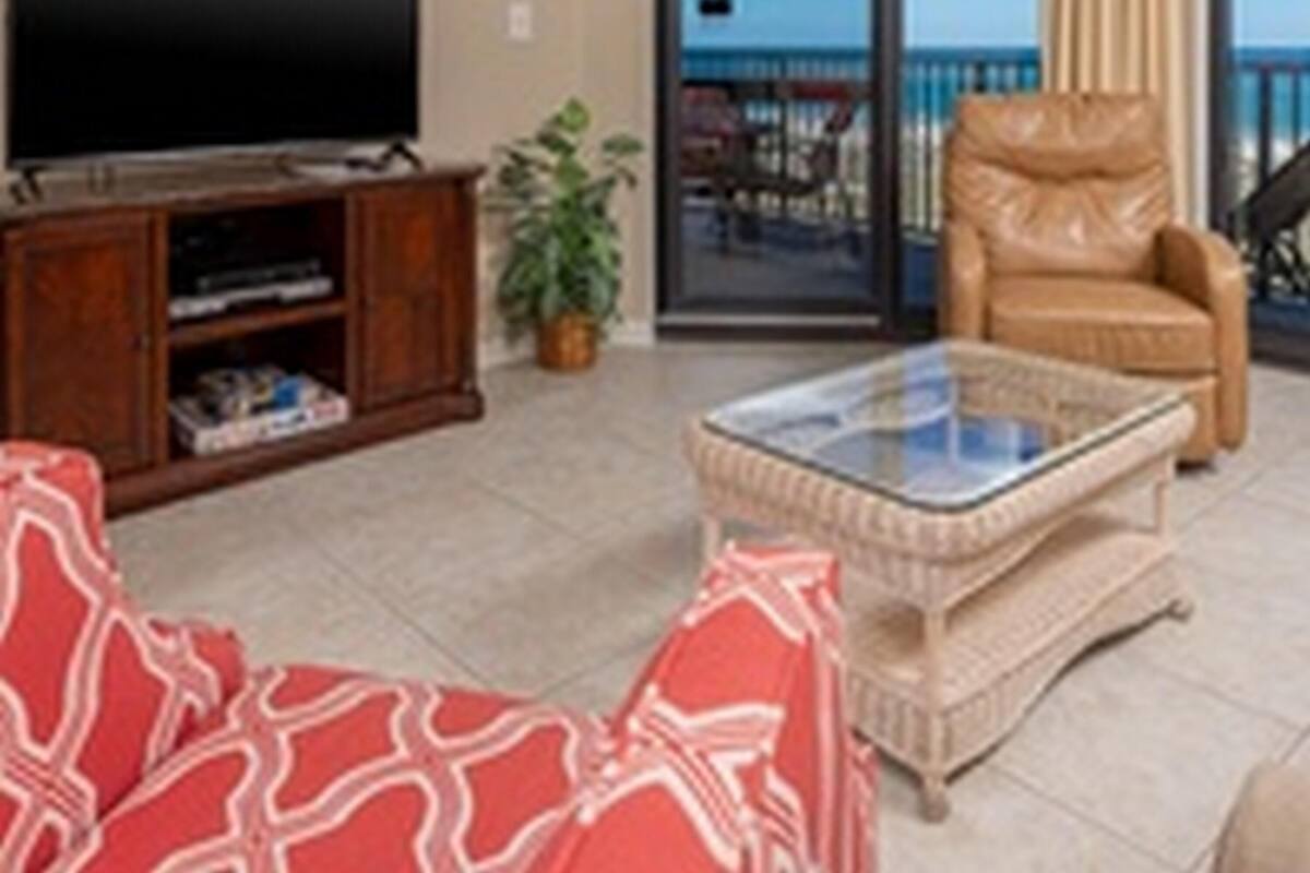 Ein Wohnzimmer mit Fernseher, einem Glastisch, einem roten, gemusterten Sofa, einem Ledersessel und Meerblick durch eine Schiebetür.