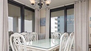 A dining area with a glass-top table surrounded by white chairs, a chandelier, and large windows with curtains. - Phoenix VI by Brett Robinson Vacations (Orange Beach)
