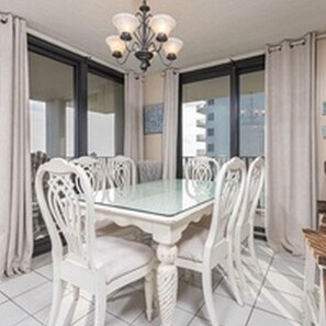 A dining area with a glass-top table surrounded by white chairs, a chandelier, and large windows with curtains.