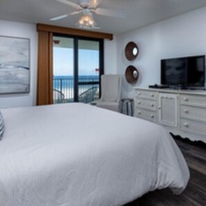 A bedroom with a large bed, a ceiling fan, a TV, and a view of the ocean through a sliding door.