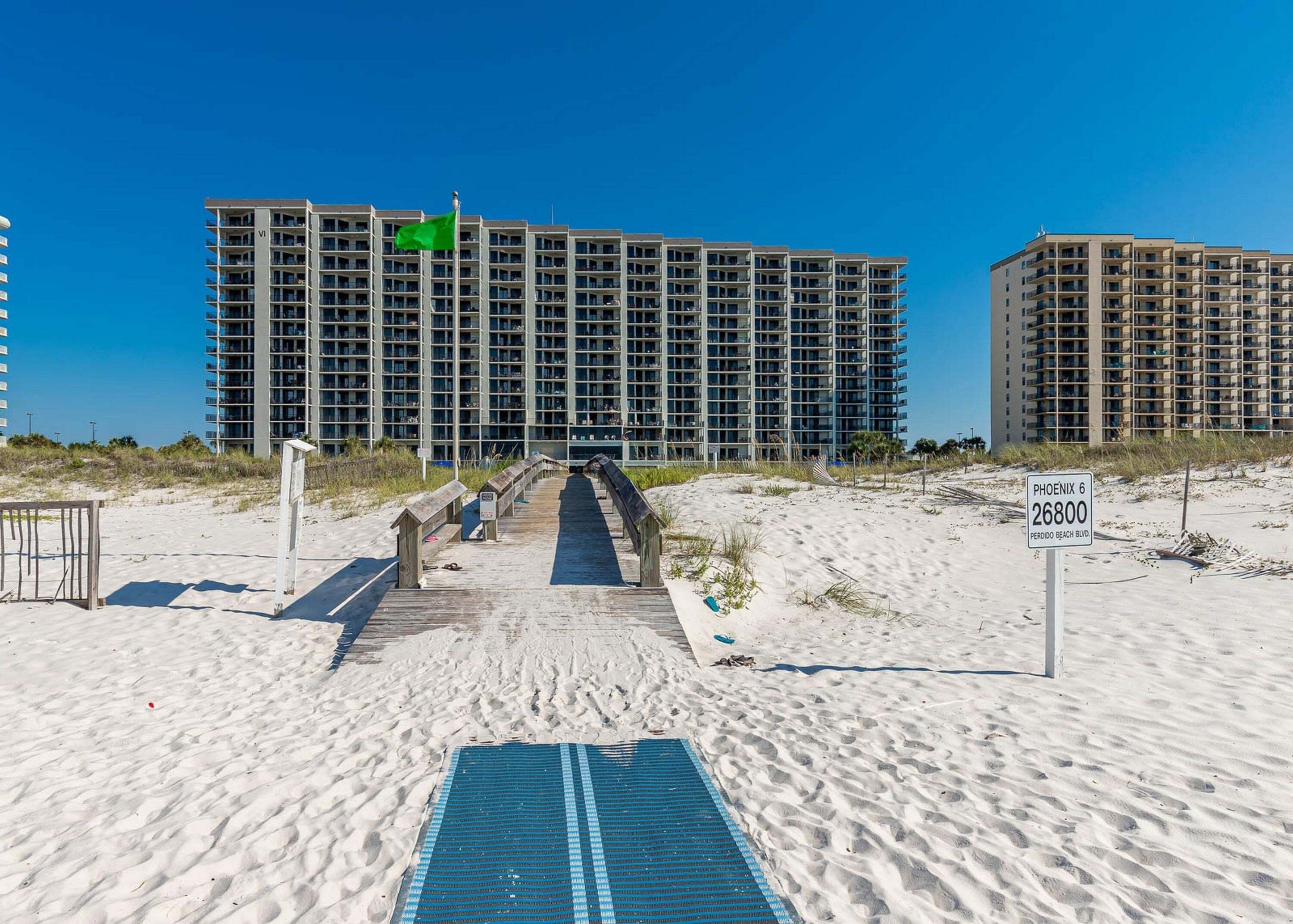 Un hotel frente al mar con un paseo marítimo que conduce a la entrada, un letrero que indica 'PHOENIX 6 26800 PERDIDO BEACH BLVD.' y un cielo azul despejado.