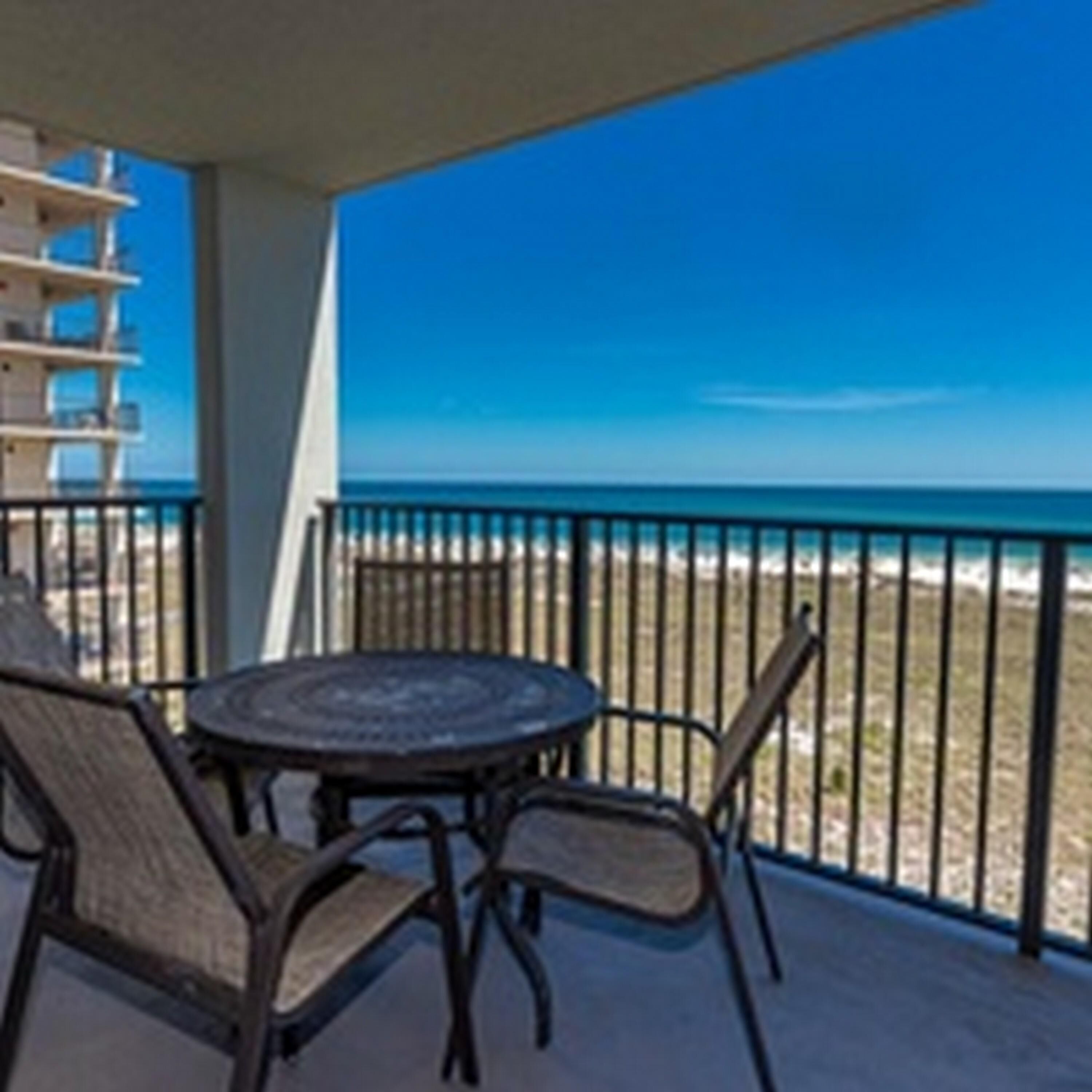 A balcony with a round table and chairs, overlooking a beach with a clear blue sky.