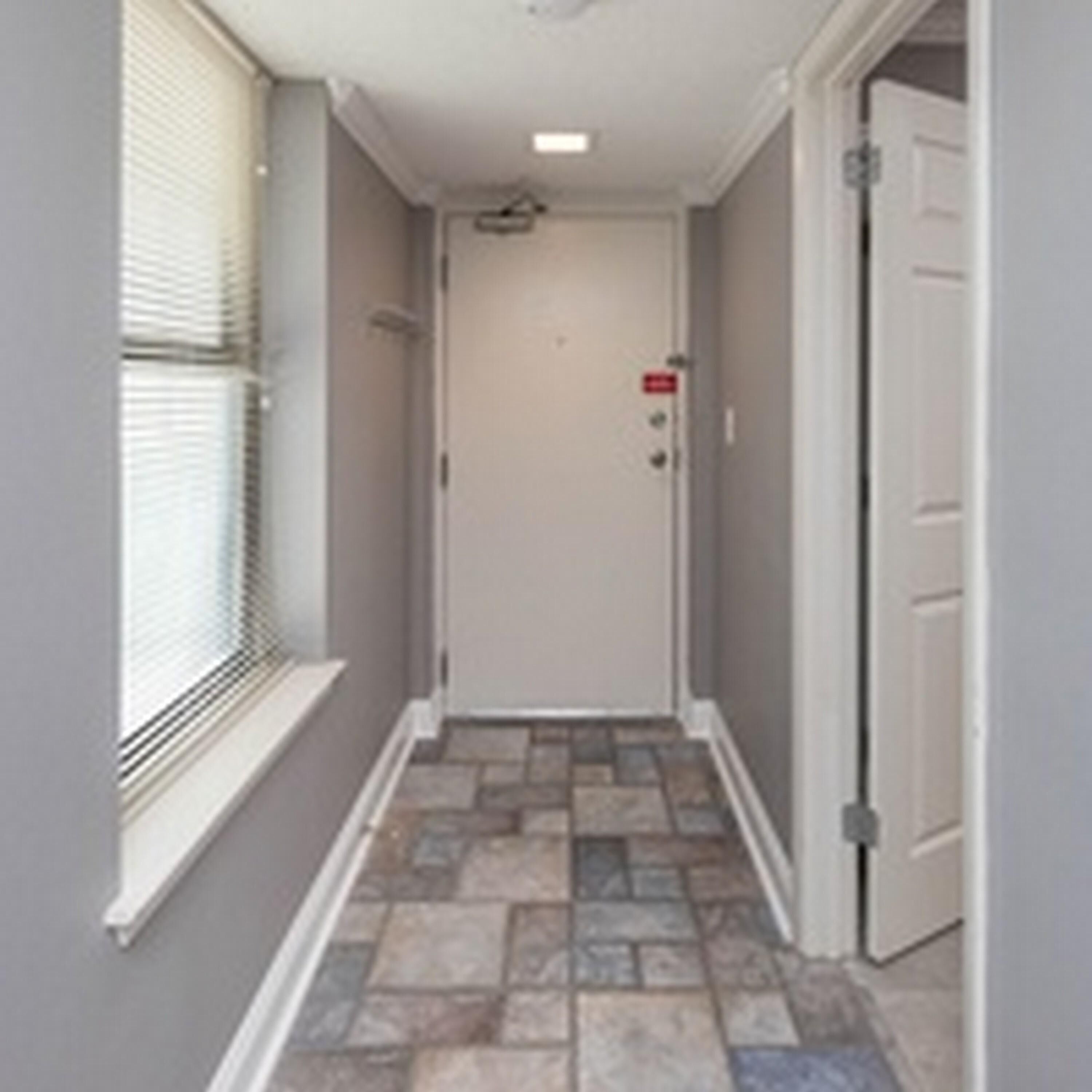A narrow hallway with a tiled floor, a door with a red emergency exit sign, and a window with blinds.