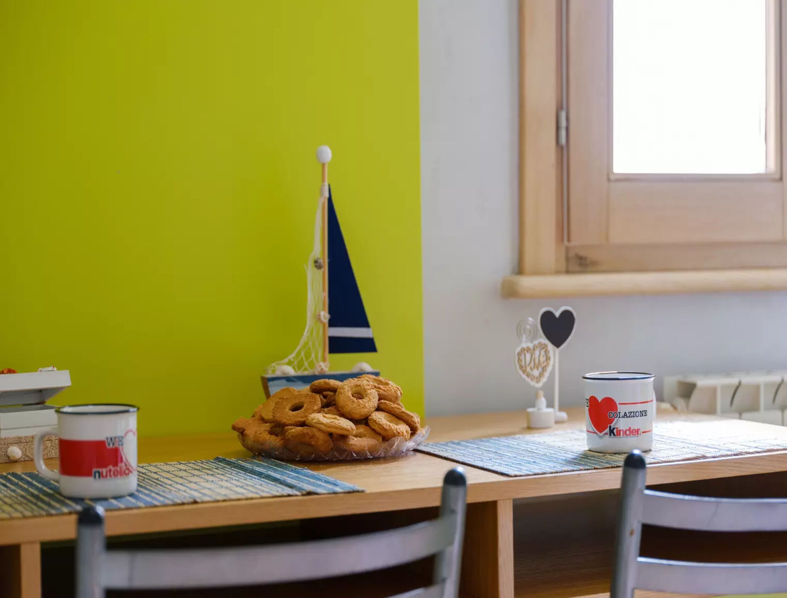 Une table à manger avec une assiette de biscuits, une petite décoration représentant un voilier, un ornement en forme de cœur et une boîte de chocolats Kinder.