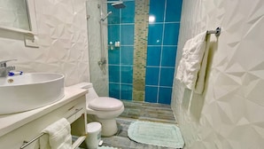 Un baño con lavamanos, inodoro y ducha blancos. Hay dos toallas en el suelo.
