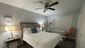 Ein Schlafzimmer mit einem Bett, Nachttischen, einem Schreibtisch, einem orangefarbenen Sessel, einem Deckenventilator und einem Fenster mit Jalousien.