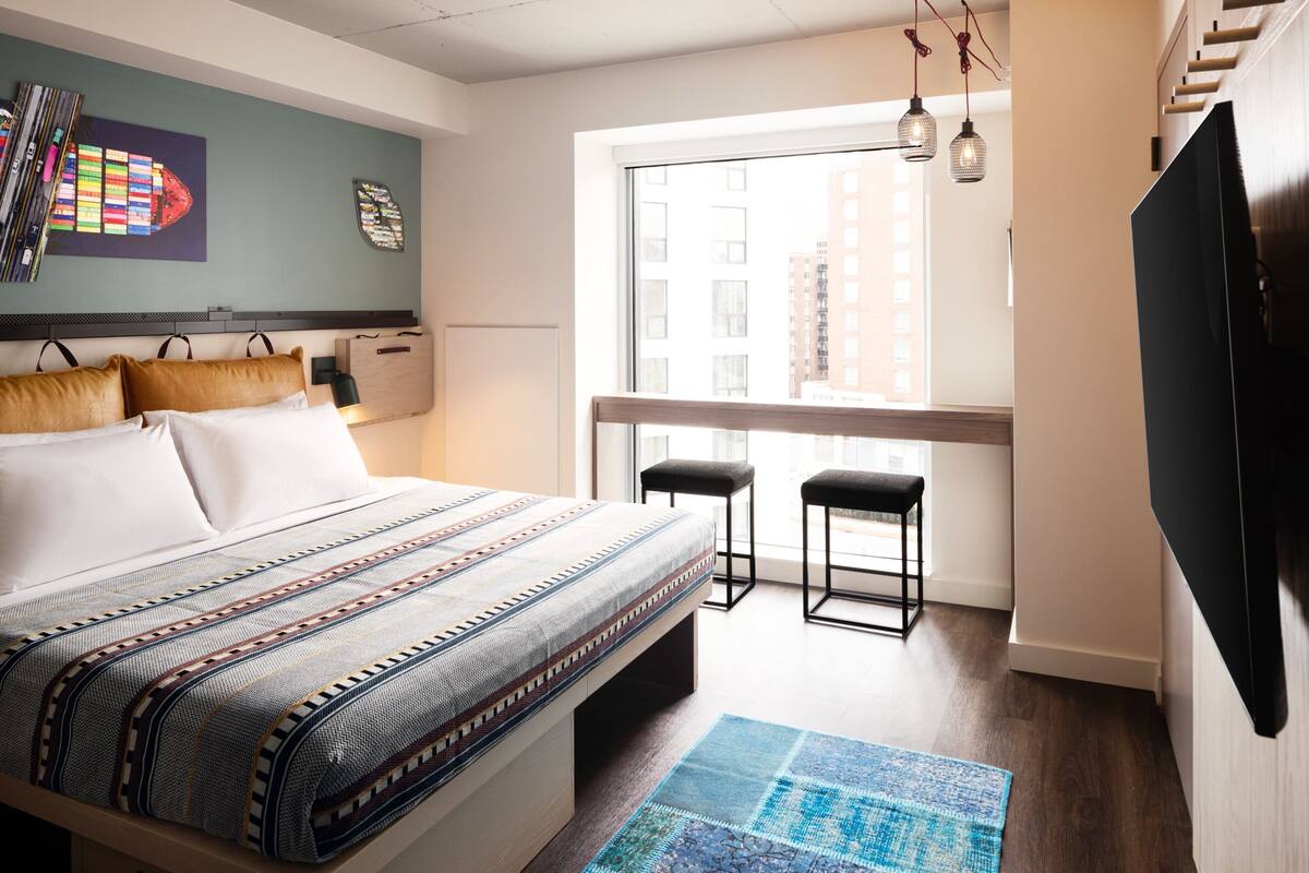 A modern bedroom with a large bed, two black stools, a TV, and a window with a city view.
