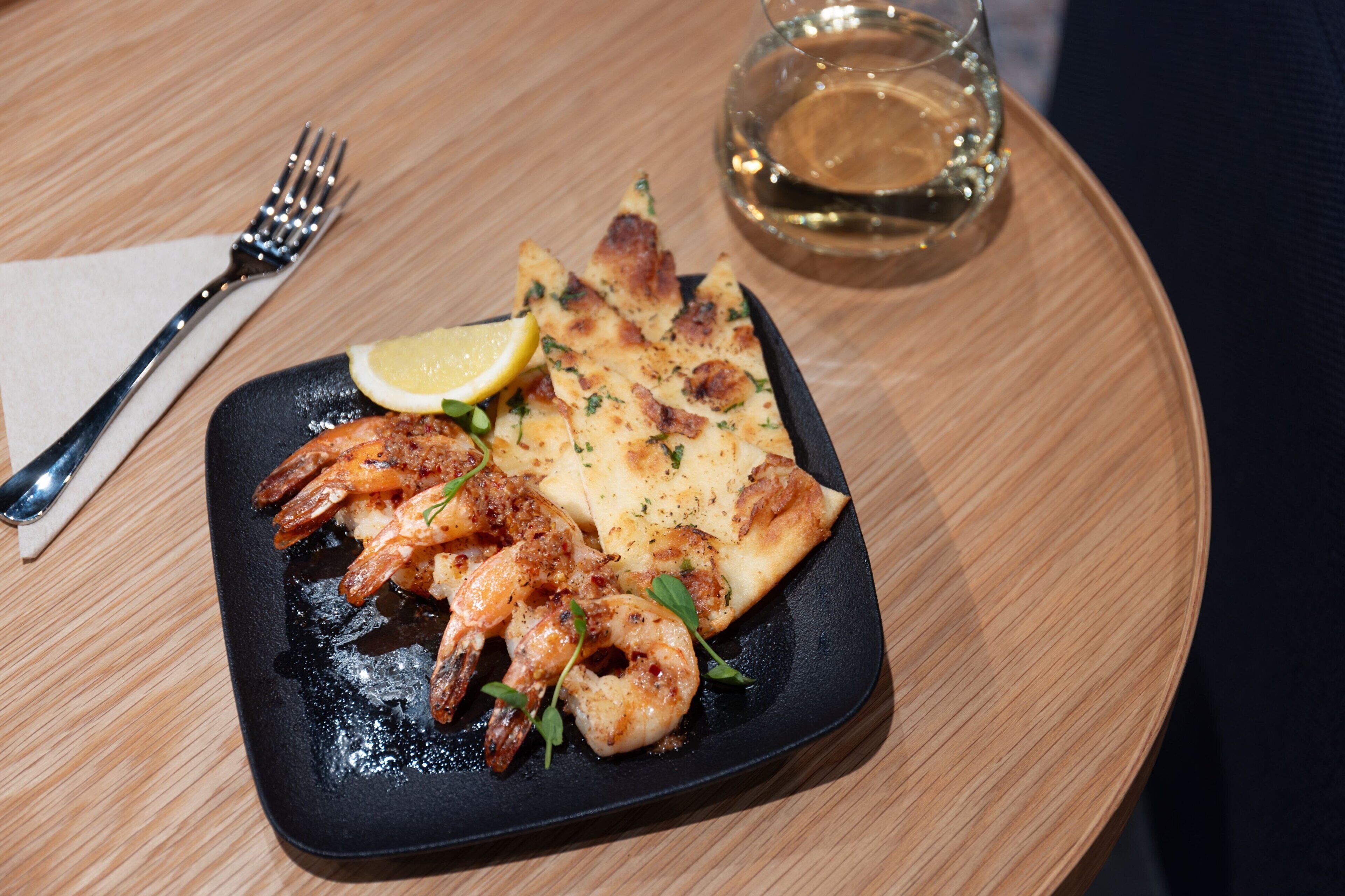 Un plato de camarones a la plancha y flatbread sobre una mesa de madera, con un tenedor y un vaso de agua.