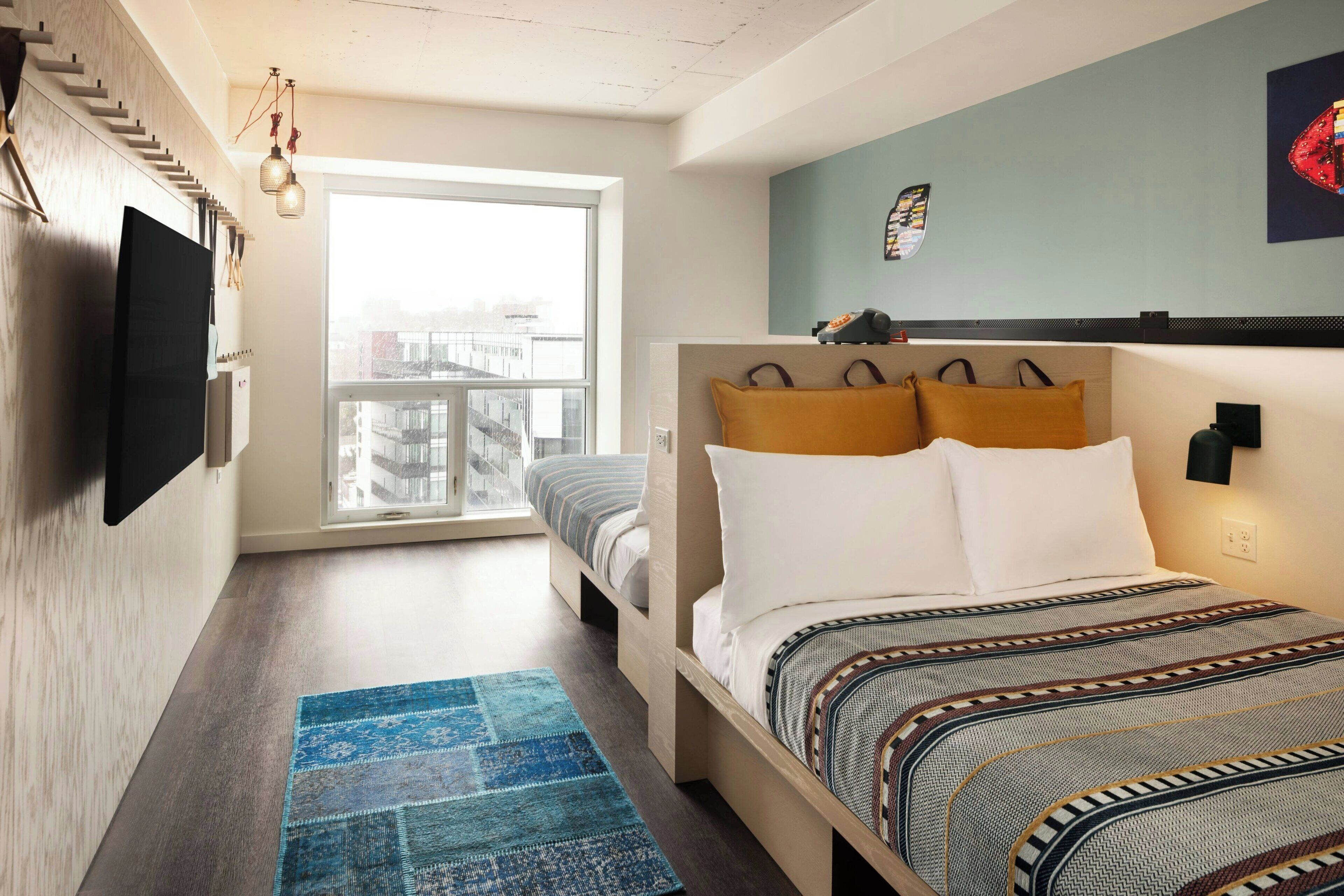 Un dormitorio moderno con una cama grande, un televisor de pantalla plana en la pared, una ventana con vista a la ciudad y una alfombra azul estampada.