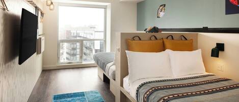 Un dormitorio moderno con una cama grande, un televisor de pantalla plana en la pared, una ventana con vista a la ciudad y una alfombra azul estampada.