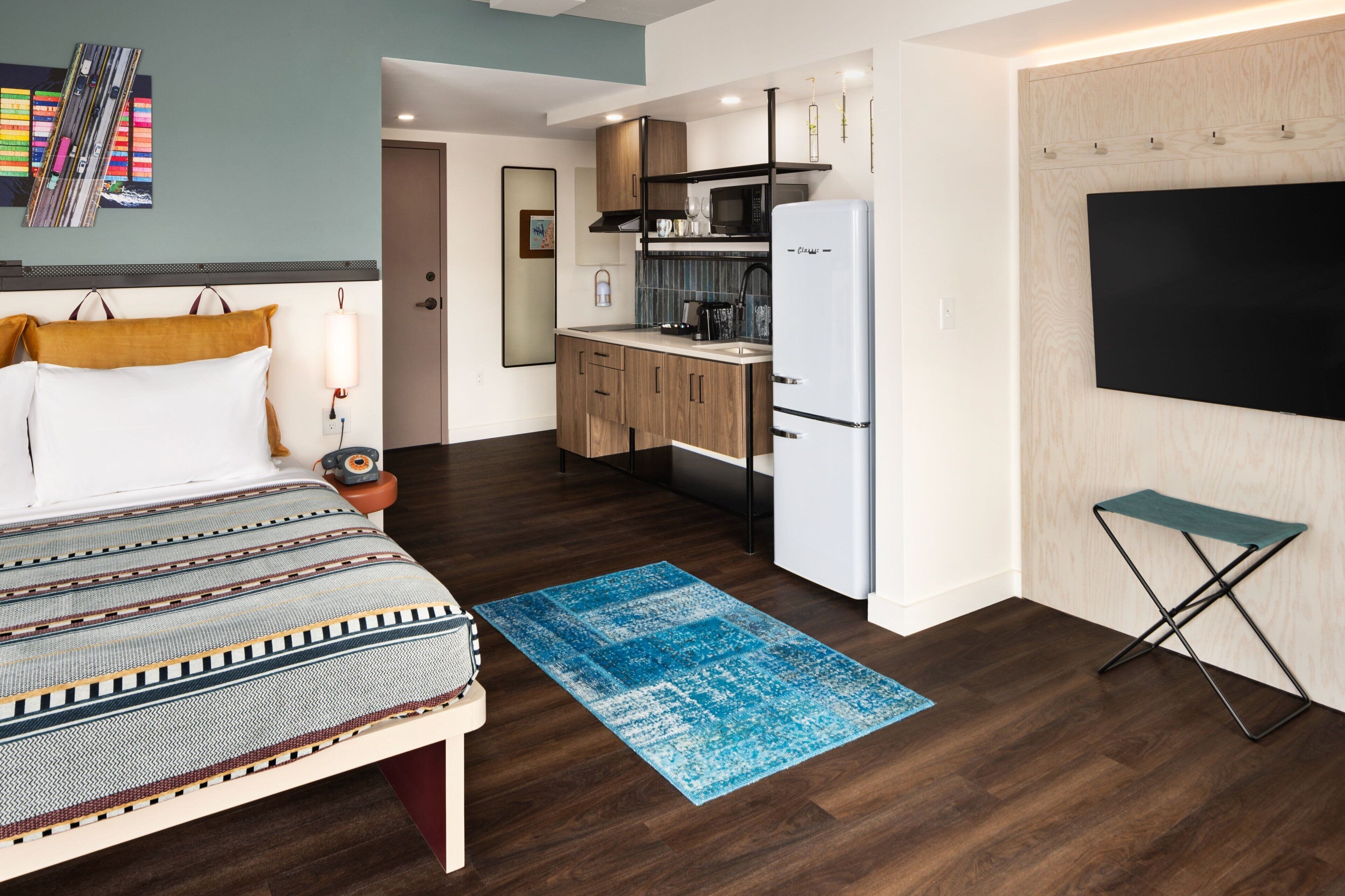 Une chambre d’hôtel moderne équipée d’un lit, d’une petite kitchenette, d’un téléviseur à écran plat et d’un tapis bleu.