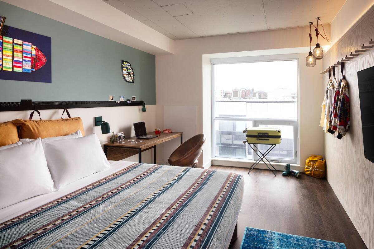 A modern hotel room with a large bed, a desk, a laptop, and a view of the city through a large window.