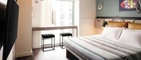 A hotel room with a large bed, a TV, a window with a view of buildings, and a small seating area with stools.