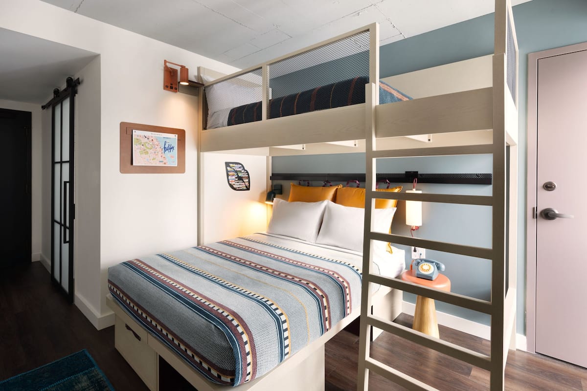 A bunk bed with a patterned bedspread, a small table with a lamp, and a door to the right.