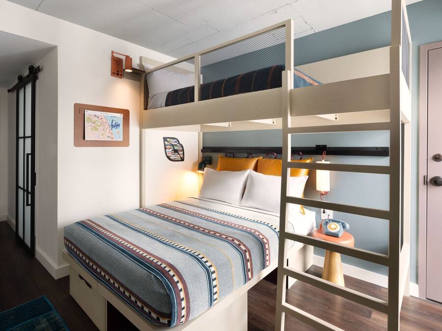 A bunk bed with a patterned bedspread, a small table with a lamp, and a door to the right.