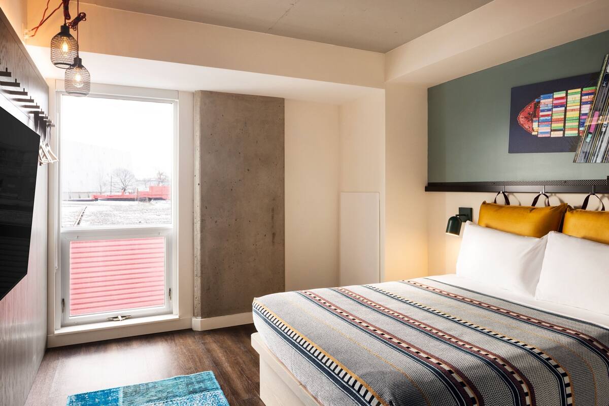 A bedroom with a large bed, a window with blinds, a hanging light fixture, and a wall-mounted TV.